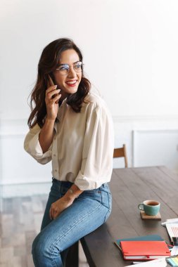 Kafede kapalı bir masada otururken cep telefonuyla konuşan gülümseyen genç bir kadın.