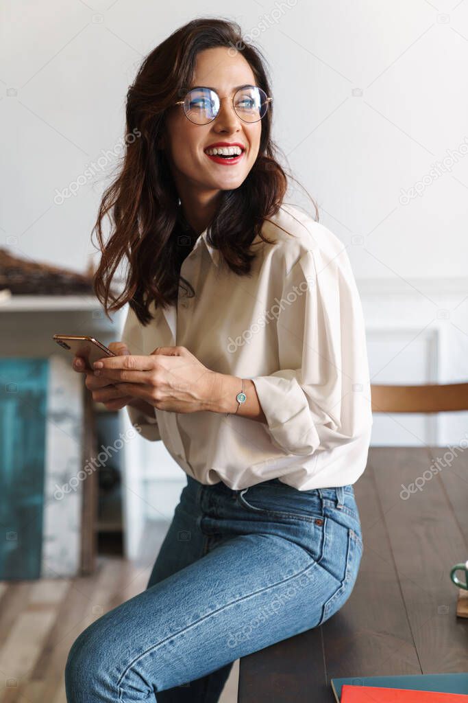 Smiling young woman using mobile phone while sitting on a table in the cafe indoors