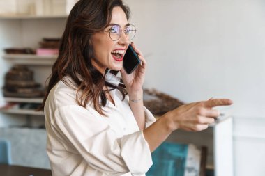 Kapalı bir kafede cep telefonuyla konuşan gülümseyen genç bir kadının resmi.