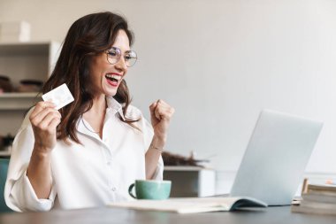 Çekici, neşeli, esmer iş kadını kafeterya masasında dizüstü bilgisayarla oturuyor, kredi kartıyla internetten alışveriş yapıyor.