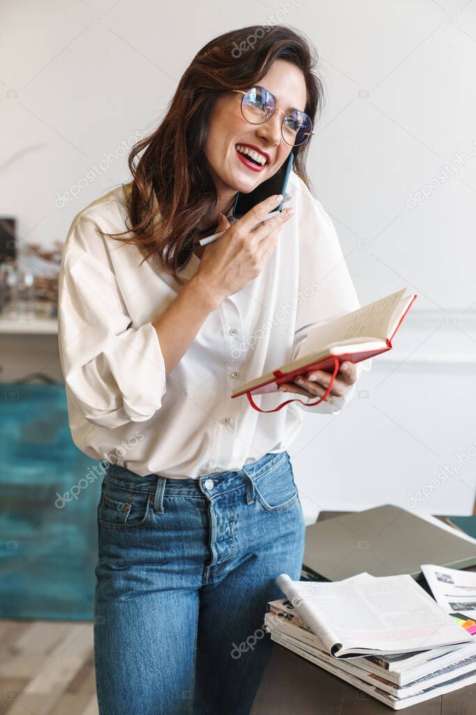 Beautiful smiling young brunette woman talking on mobile phone while studying in the cafe indoors, standing at the table