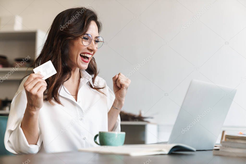 Attractive cheerful young brunette businesswoman sitting at the cafe table with laptop computer indoors, shopping online with credit card