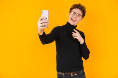 Selfie çeken ve sarı arka planda izole edilmiş cep telefonunu işaret eden gülümseyen genç bir adam.