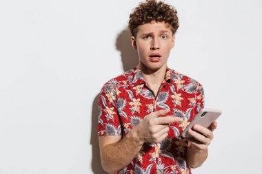 Kafası karışmış genç bir adamın beyaz arka planda izole edilmiş cep telefonunu kullanması ve işaret etmesi