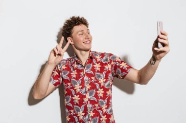 Cep telefonuyla selfie çeken ve beyaz arka planda izole edilmiş barış işareti yapan genç bir adam görüntüsü