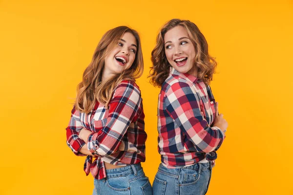 两个穿着格子呢衬衫的年轻漂亮姑娘笑着背靠背站在黄色背景上的照片 — 图库照片