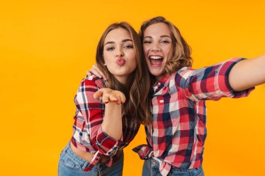 Sarı arka planda izole edilmiş selfie çekilirken gülümseyen ve kucaklaşan ekose gömlekli iki güzel kızın resmi