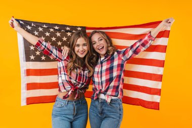 Ekose gömlek giyen, gülümseyen iki güzel kız ve sarı arka planda izole edilmiş Amerikan bayrağı.