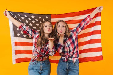 Ekose gömlek giyen, gülümseyen iki güzel kız ve sarı arka planda izole edilmiş Amerikan bayrağı.