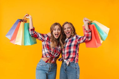 Ekose gömlek giyen, gülümseyen ve sarı arka planda izole edilmiş renkli alışveriş torbaları taşıyan iki güzel kız resmi.