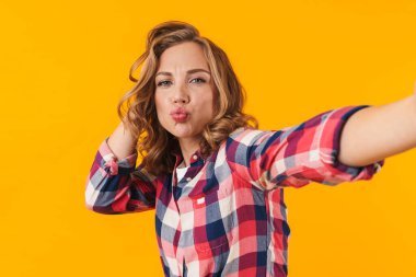 Ekose gömlek giyen, gülümseyen ve sarı arka planda izole edilmiş selfie çeken güzel bir kadın resmi.