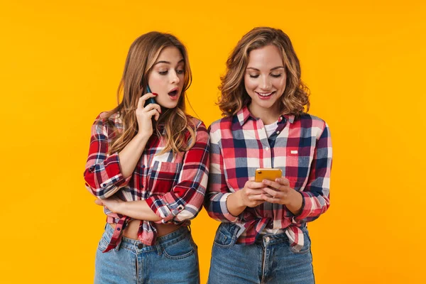 再生シャツを着て笑顔と黄色の背景に隔離された携帯電話を使用して2人の若い美しい女の子のイメージ — ストック写真