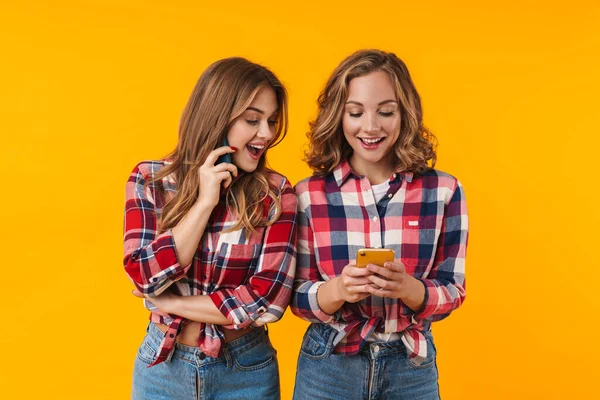 再生シャツを着て笑顔と黄色の背景に隔離された携帯電話を使用して2人の若い美しい女の子のイメージ — ストック写真