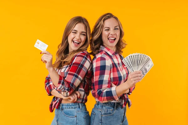 两个年轻貌美的女孩穿着格子呢衬衫 手里拿着美元钞票和信用卡 背景是黄色的 — 图库照片