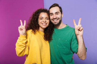 Mor arka planda izole edilmiş barış parmaklarını gösterirken renkli giysiler içinde gülümseyen Kafkas çift portresi.