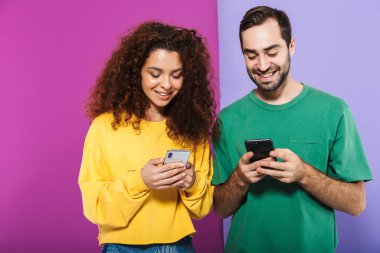 Renkli giyinmiş, gülümseyen ve cep telefonlarını bir arada tutan beyaz bir çiftin portresi menekşe arka planda izole edilmiş.