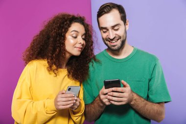Neşeli Kafkas çift portresi. Renkli giysiler içinde, gülümseyen ve cep telefonlarını bir arada tutan kadın ve erkek. Menekşe arka planda izole edilmiş.