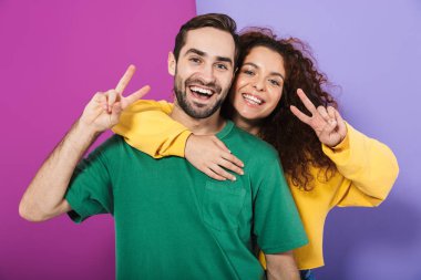 Renkli elbiseli mutlu beyaz çift portresi. Mor arka planda izole edilmiş barış parmaklarını gösterirken sarılıyorlar.