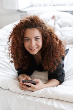 İç çamaşırı ipek bornozlu, mutlu, genç bir kadının görüntüsü evde, yatağında cep telefonu kullanarak yatıyor..