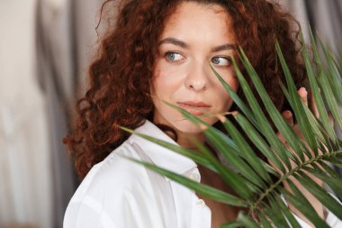 Kızıl saçlı, iç çamaşırlı, yeşil yaprak bitkisinin yanında beyaz gömlekli bir kadın tasviri..