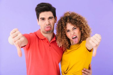 Genç hoşnutsuz bir çiftin portresi dillerini uzatan ve baş parmaklarını gösteren mor arka planda izole edilmiş.