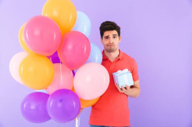 Kafası karışmış bir adamın görüntüsü. Elinde çok renkli balonlar ve menekşe arka planda izole edilmiş hediye kutusuyla dikilirken kaşlarını çatıyor.