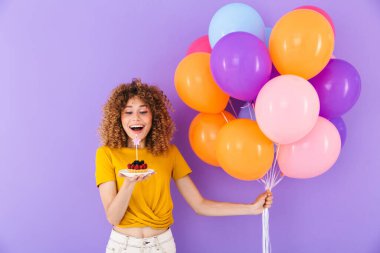 Mutlu genç bir kadının doğum gününü çok renkli balonlarla ve menekşe arka planda izole edilmiş bir parça turtayla kutlaması.