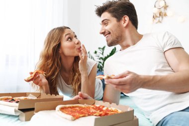 Evdeki yatakta uzanmış pizza yiyen mutlu bir çiftin resmi.