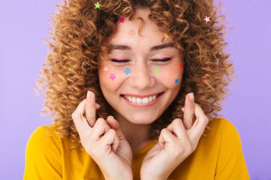 İyimser, kıvırcık bir kızın, yüzünde mor arkaplan üzerinde izole edilmiş çok renkli parıldayan yıldızlarla parmaklarını çapraz yaptığı yakın plan görüntüsü.