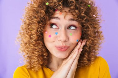 Güzel, kıvırcık bir kızın görüntüsü. Gülümseyen ve yüzünde mor arkaplanda izole edilmiş çok renkli parıldayan yıldızlarla hayaller kuran.