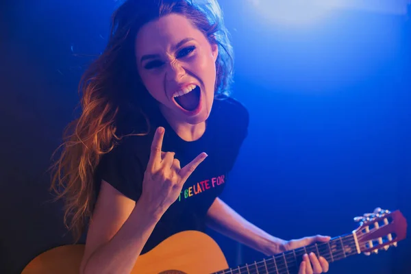 Image of emotional amazing young positive woman musician play on a guitar on scene in night club over dark background with flash lights showing rock gesture.