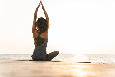 Eşofmanlı esmer sporcu kadının iskelede oturduğu ve sabahları deniz kenarında yoga yaptığı fotoğraf.