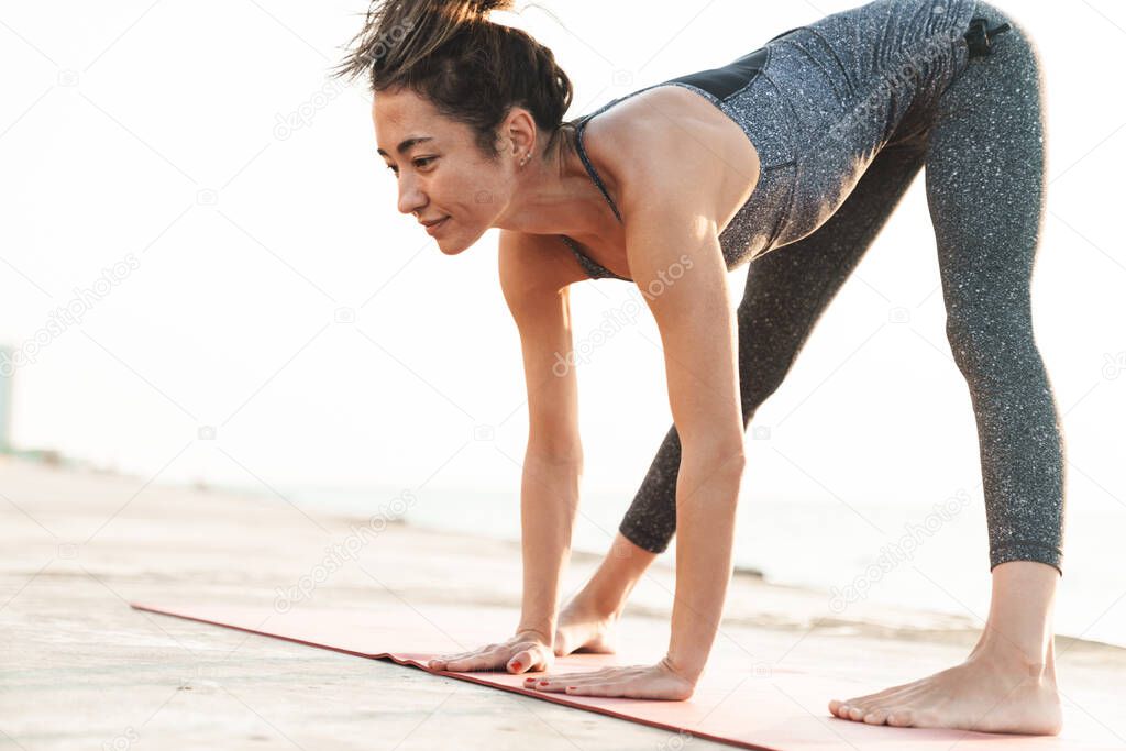 Photo of beautiful sportswoman in tracksuit stretching her body on mat while practicing yoga by seaside in morning