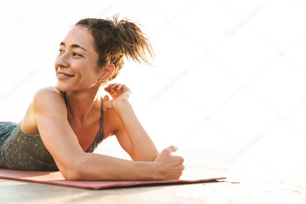 Photo of caucasian sportswoman in tracksuit resting and lying on fitness mat while practicing yoga by seaside in morning