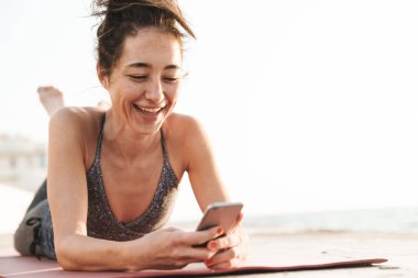 Eşofmanlı gülümseyen sporcu kadının elinde akıllı telefon tutarken ve sabahları deniz kenarında yoga yaparken spor minderinde yatarken fotoğrafı.
