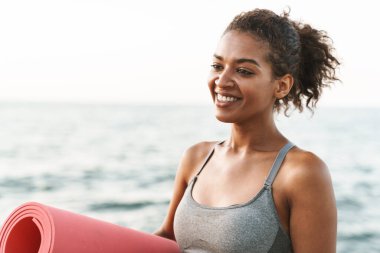 Eşofmanlı neşeli Afro-Amerikan sporcu kadının fotoğrafı. Sabahları deniz kenarında yoga minderiyle gülümsüyor ve ayakta duruyor.