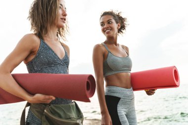 Eşofman giymiş, gülümseyen ve konuşan iki güzel spor kadının fotoğrafı. Sabahları deniz kenarında yoga minderleriyle dikilirken.