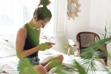 Parlak odada yeşil çiçekli dergi okurken elinde cep telefonu olan neşeli kadın portresi.