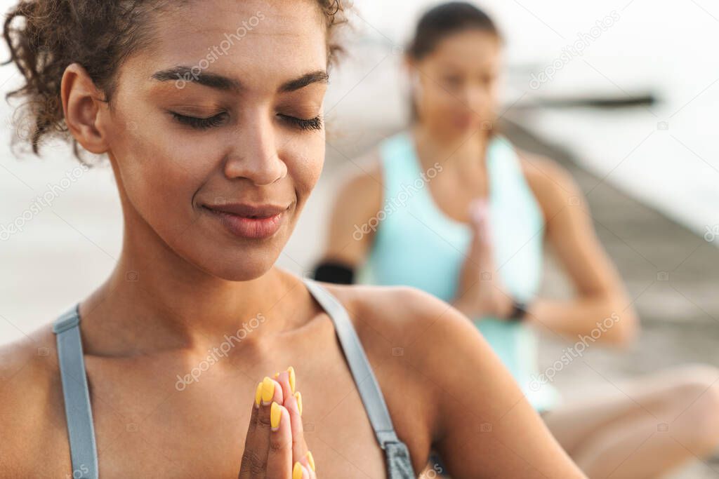 Photo of calm multiethnic sportswomen in tracksuits meditating and keeping palms together while sitting on yoga mats by seaside in morning