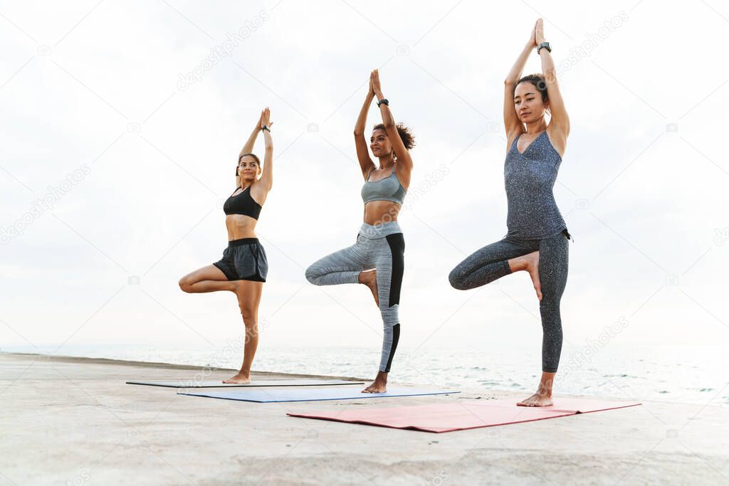 Photo of pleased multiethnic sportswomen in tracksuits doing exercises with palms together on yoga mats by seaside in morning