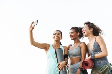 Eşofmanlı, neşeli, çoklu etnik kökenli üç kadın dışarıda beyaz duvarın üzerinde dururken gülümseyen ve cep telefonuyla selfie çeken.