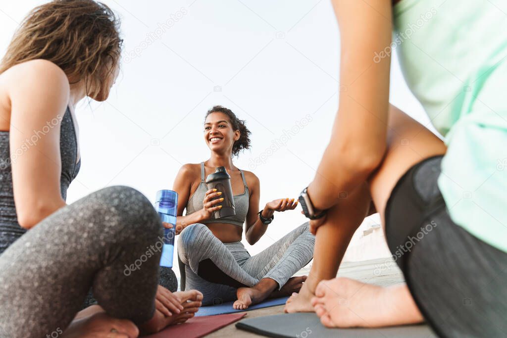 Photo of cheerful multiethnic sportswomen in tracksuits smiling and talking while sitting on yoga mats by seaside in morning