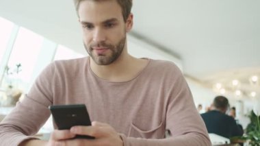 Memnun genç sakallı bir adam kafeteryada otururken akıllı telefonunu kullanıyor.