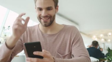 Mutlu, genç, sakallı bir adam kafeteryada otururken akıllı telefonunu kullanıyor.