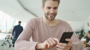 Memnun genç sakallı bir adam kafeteryada otururken akıllı telefonuyla yazı yazıyor.