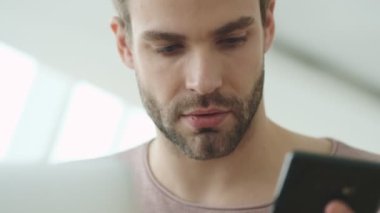 Çekici genç sakallı bir adamın yakın plan görüntüsü bilgisayarına bakıyor ve kafeteryada otururken akıllı telefonuyla konuşuyor.