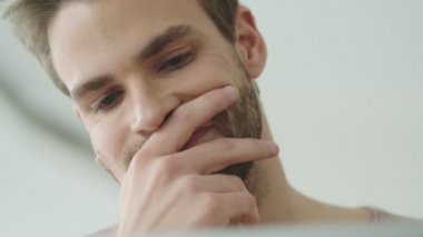 Odaklanmış sakallı genç bir adamın yakın plan görüntüsü bilgisayarına bakıyor ve kafede otururken yüzüne dokunuyor.