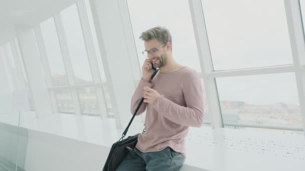 一位面带微笑 头戴眼镜 身穿休闲装的年轻人站在一家白色公共场所的窗户边 一边用智能手机聊天 — 图库视频影像
