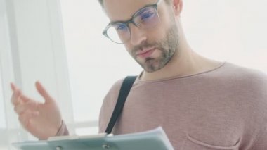 Kafası karışmış, gözlüklü genç bir adam pencerenin yanında durup belgeleri okuyor.
