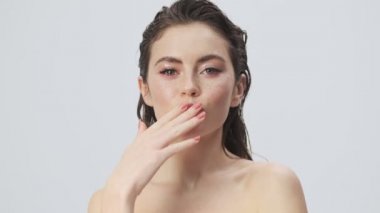 Göz kamaştırıcı, yarı çıplak, ıslak saçlı bir kadın stüdyoda açık mavi bir arkaplanda kameraya hava öpücüğü gönderiyor.